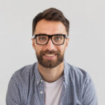 smiling man with a beard and black-framed glasses wearing a blue checked shirt over a white t-shirt