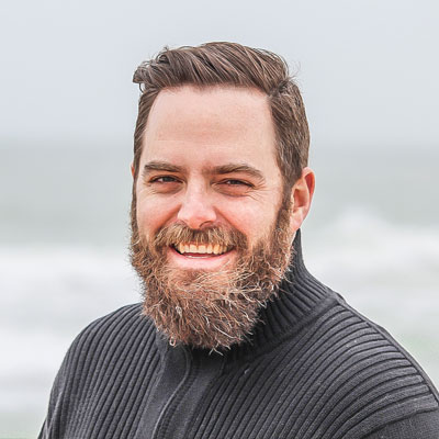 man with a groomed beard and brown hair smiling outdoors, wearing a black textured sweater