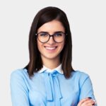 smiling young woman with dark hair and black-framed glasses wearing a blue pussy-bow blouse on a white background