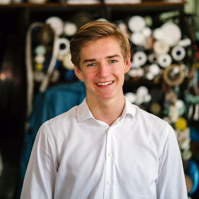 A young man with light brown hair and a white button-down shirt smiling in front of a blurred background