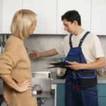plumber in blue overalls talking to a female homeowner while holding a digital tablet in a modern kitchen