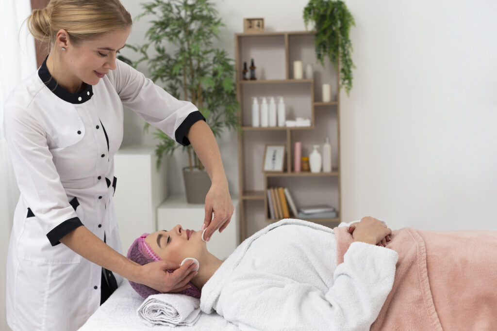 smiling female aesthetician in a white uniform performing a facial treatment on a client in a spa