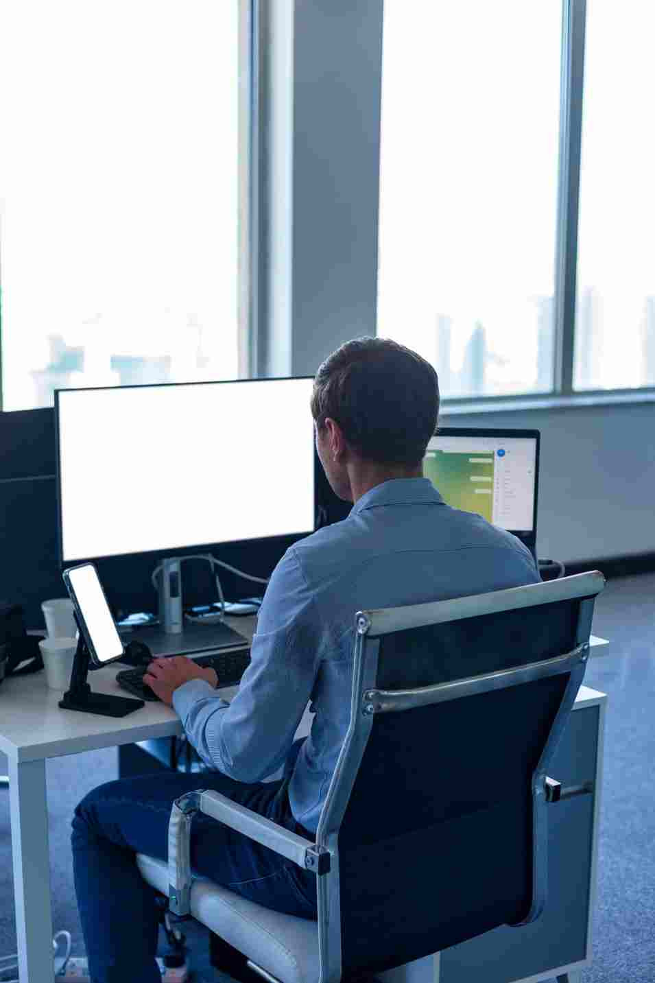Man in a blue shirt working at a desk with multiple monitors and a smartphone in a bright, modern corporate office