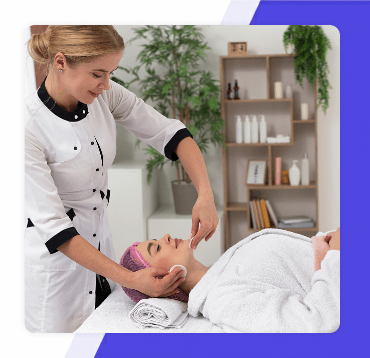 smiling female aesthetician in a white uniform performing a facial treatment on a client in a spa