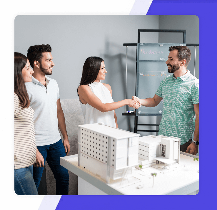 A woman in a white top shaking hands with a man over an architectural model while a young couple looks on
