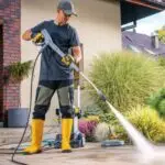 worker in yellow boots and a cap using a high-pressure washer to clean a stone patio outside a house