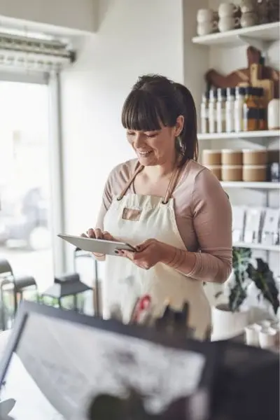 smiling female shop owner in a beige apron using a tablet in her brightly lit boutique