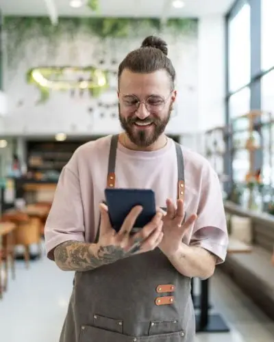 smiling male barista with a man bun and tattoos using a tablet in a modern, sunlit cafe