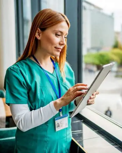 female nurse in teal scrubs and a lanyard using a digital tablet while standing by a large window