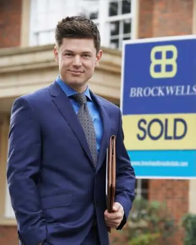 male real estate agent in a blue suit standing in front of a blue and yellow "SOLD" sign