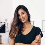 smiling female hairstylist in a black t-shirt, holding professional shears with her arms crossed in a bright salon