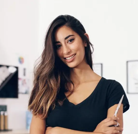 smiling female hairstylist in a black t-shirt, holding professional shears with her arms crossed in a bright salon